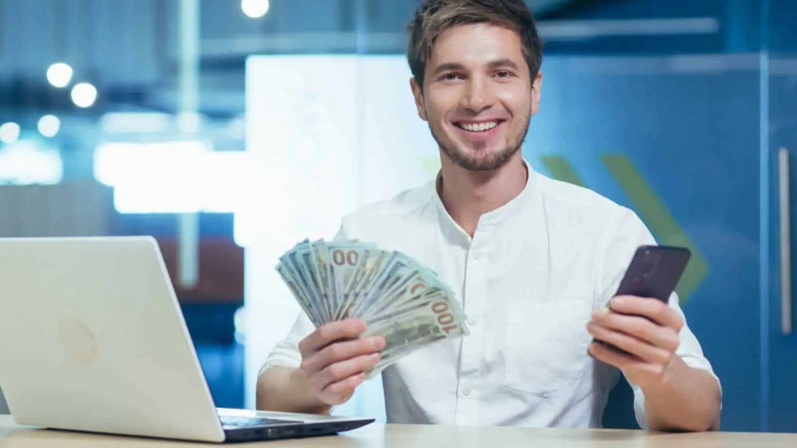 Happy and satisfied young handsome man. Businessman holding a wad of money and a mobile phone. Sits in the office at the desk, shows, brags. smiling