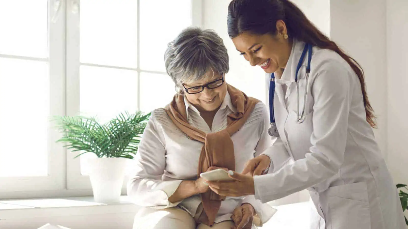 Caring doctor teaches female patient to use mobile healthcare app. Retired lady sitting in hospital exam room looking at cell screen learning to download health tracker for senior citizens. Copy space