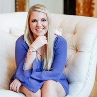 Woman with blonde hair and a purple dress sintting on a white, high back couch chair.