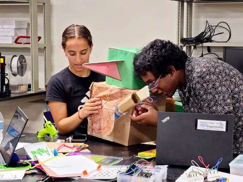 2 teenagers sitting in a classroom setting working on a cardboard box project