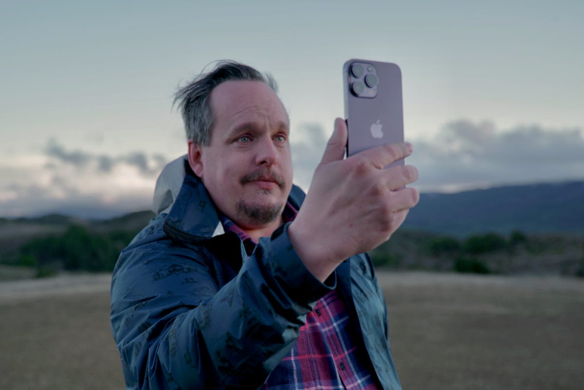 A man holding an iPhone 14 Pro outdoors on a remote hillside