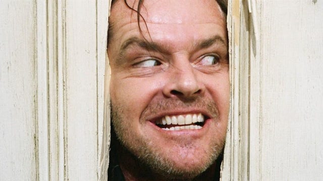 Iconic film still of Jack Nicholson as Jack Torrance in The Shining. Eyebrows raised and grinning widely, he peers sinisterly through a hole he's just bashed in a door with his axe.