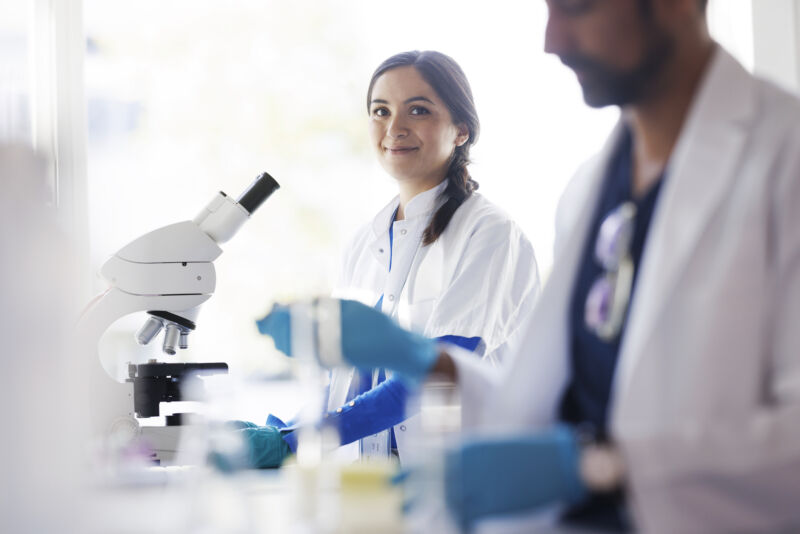 two people in lab coats wearing rubber gloves standing near a microscope.