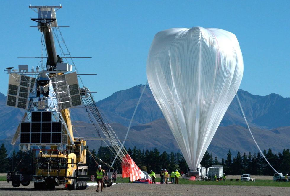 A giant white balloon being inflated outside. Next to it is a large yellow vehicle hoisting a silver foil and solar panel covered apparatus. 