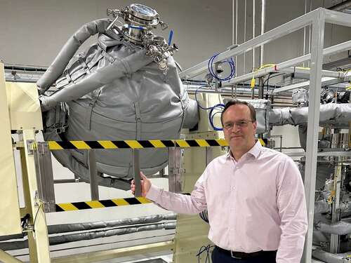 A man in glasses stands next to a large piece of equipment consisting of a rounded circle with gauges and wires.