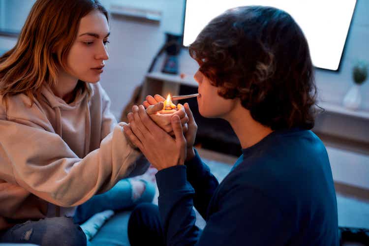 Young girl lighting cannabis cigarette for her boyfriend while sitting on the couch at home. Young couple smoking marijuana joint and relaxing