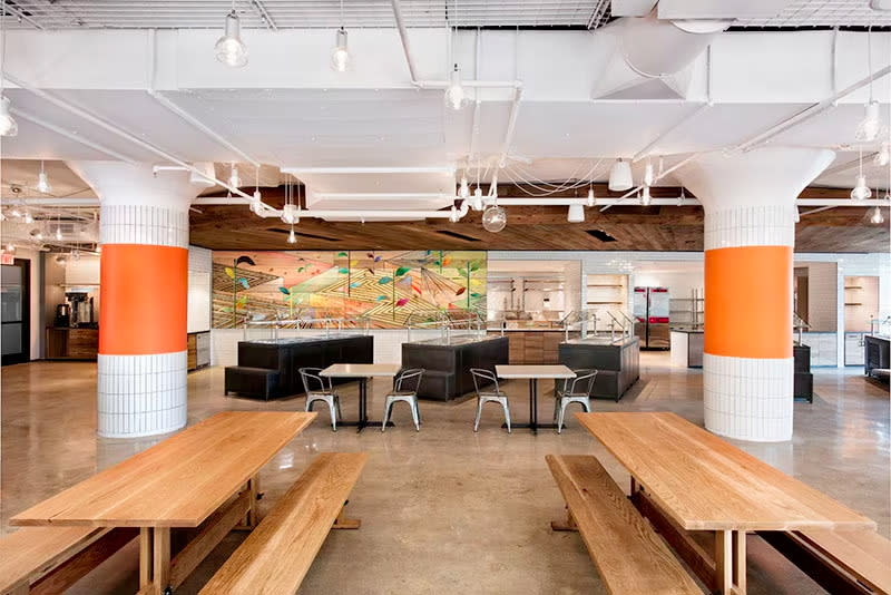 Etsy’s headquarters, inside view. A commons area includes benches and tables with pillars and buffet stations behind. Art decorates the walls.