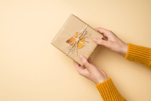 Top view photo of hands in yellow sweater giving craft paper gift box with twine bow and yellow autumn leaf.