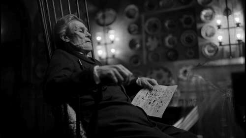 A deformed Willem Dafoe sits in a chair holding papers in a black-and-white still from Poor Things.