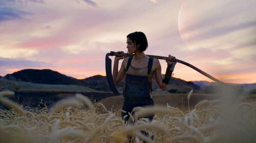 Sofia Boutella strikes a cool pose with a stick against a pretty landscape in a still from Rebel Moon – Part One: A Child of Fire