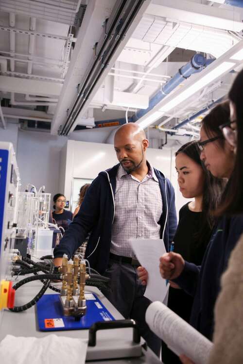 A man operates a device with hoses and metal pipes in a chemical lab while a group of students observes.