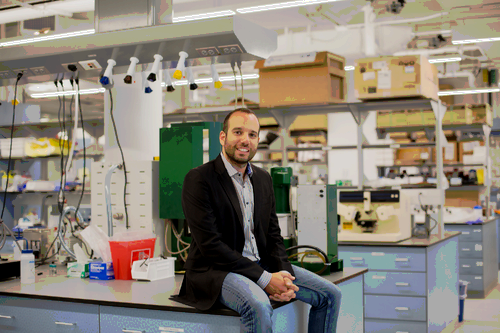 A man with brown hair, light beard, wearing jeans and dark jacket, sits on a bench in a lab with bottles, tubes, and other equipment.