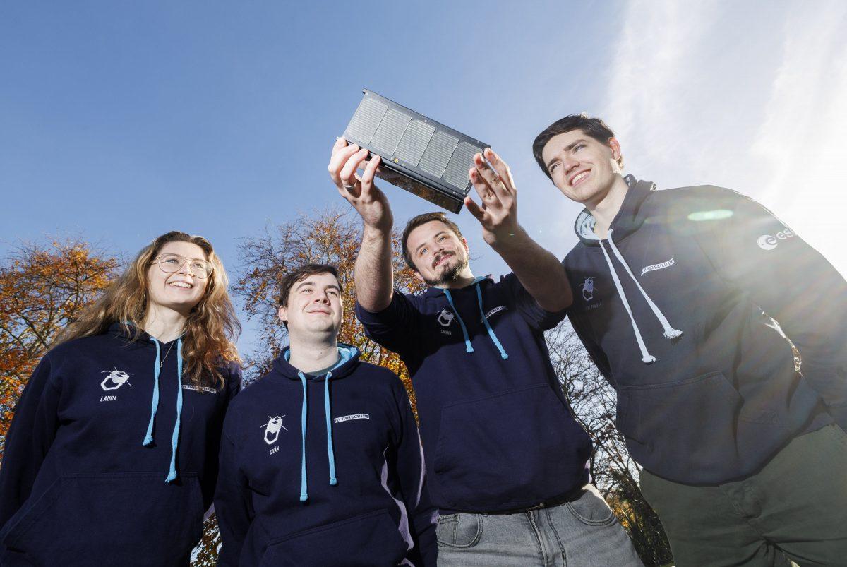 Three men and a woman standing together with a blue sky and trees in the background. One of the men is holding a small satellite in his hands, which is EIRSAT-1.