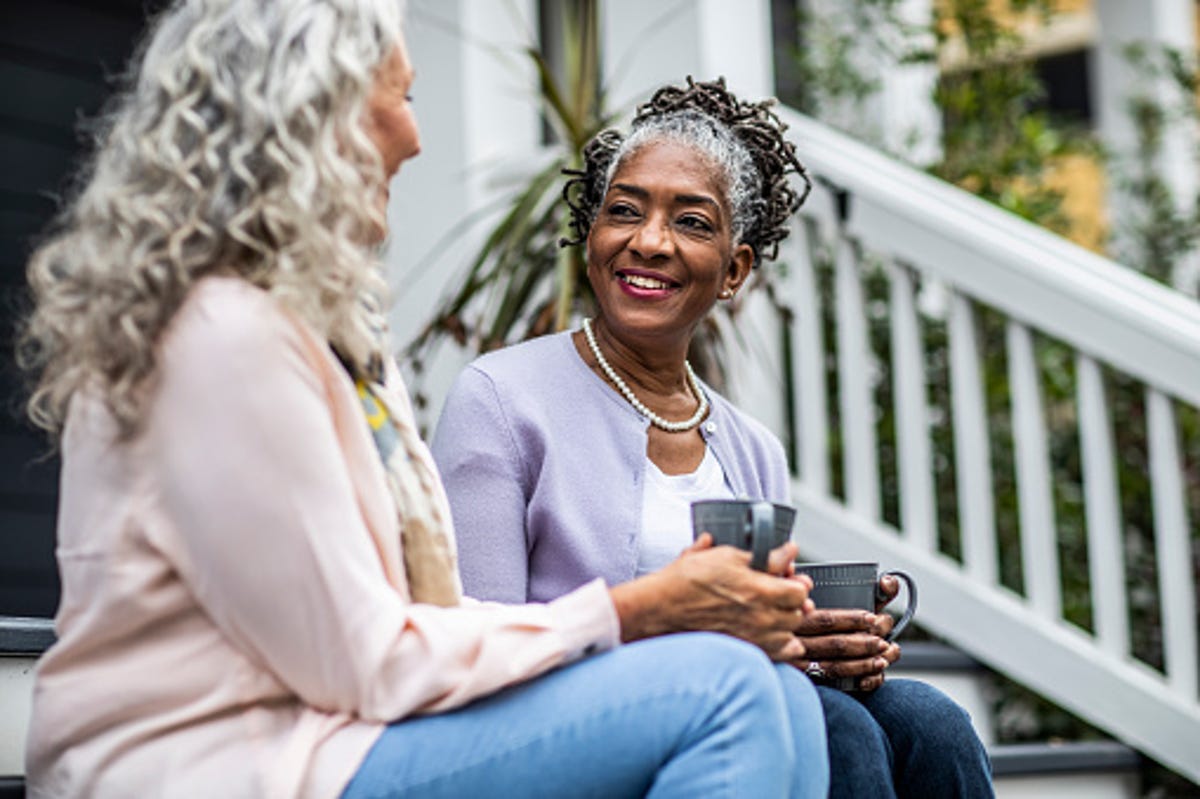 Two older women sitting on porch stairs drinking coffee in front of their house.