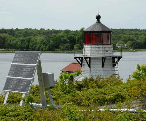 A ground mounted solar panel in front of a white lighthouse near water.