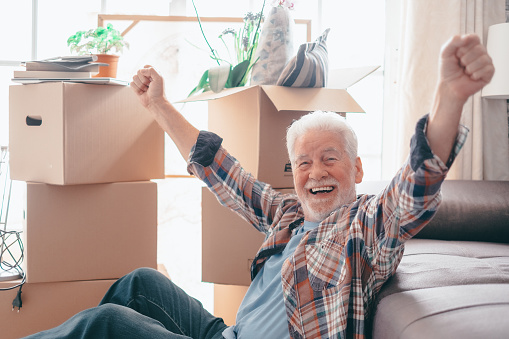 Happy senior man sitting on floor with winning gesture relaxing in new home living room with cardboard boxes packed with stuff on moving day.