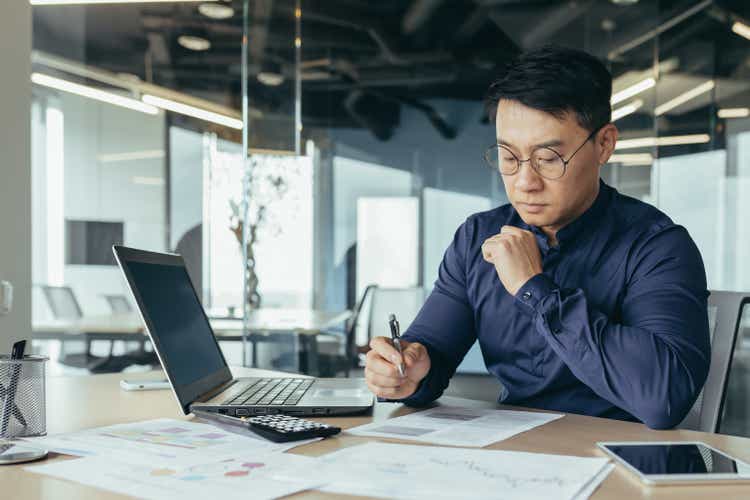 Thinking asian financier in glasses accountant working with documents and accounts, using laptop, male businessman doing paperwork in sitting at desk inside office building