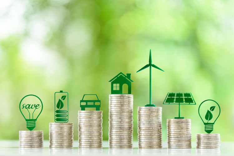 Renewable or clean energy generation prices and costs, financial concept : Green eco-friendly symbols atop coin stacks e.g. energy efficient light bulb, a battery, a solar cell panel, a wind turbine.