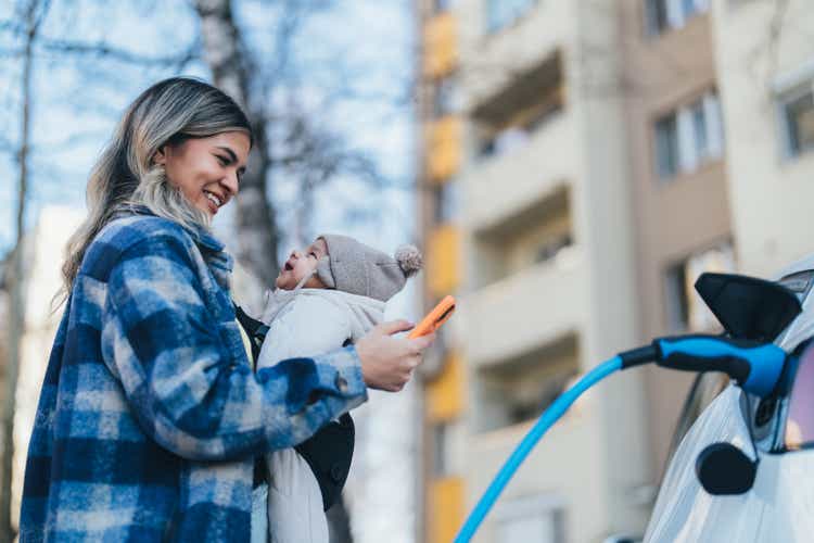 Young woman with baby charging her electric car in front of the house