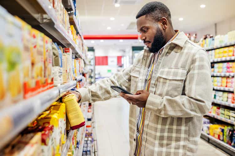 Man Looking At Smartphone While Choosing Items In Supermarket