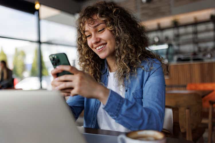 Young smiling woman using mobile phone while working on a laptop at a cafe