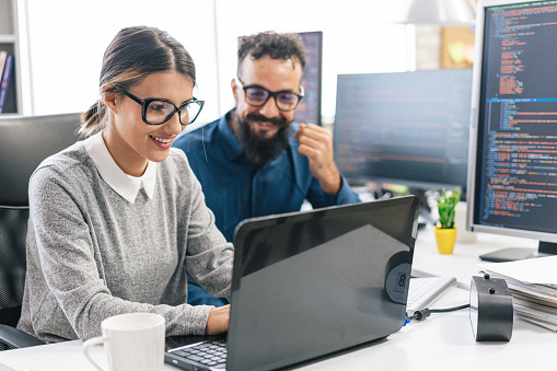 Young female programmer standing on her desk, coding on a laptop, having discussion with a male colleague, giving her advice