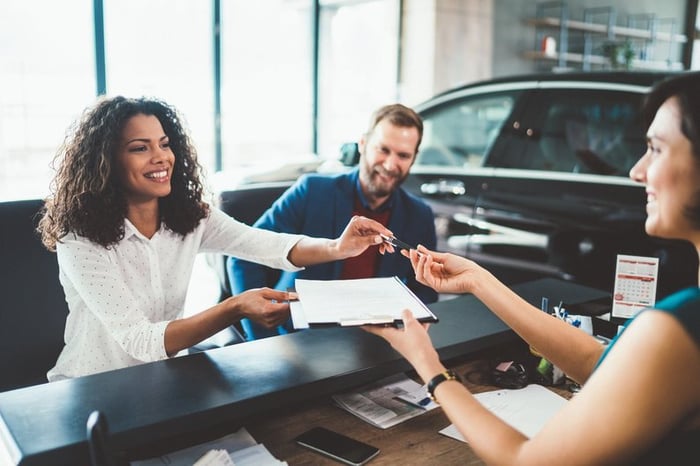 Two people sitting at a desk in a car showroom and handing signed forms back to the salesperson.