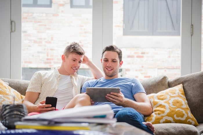 A young couple sit on a couch, looking at a tablet.