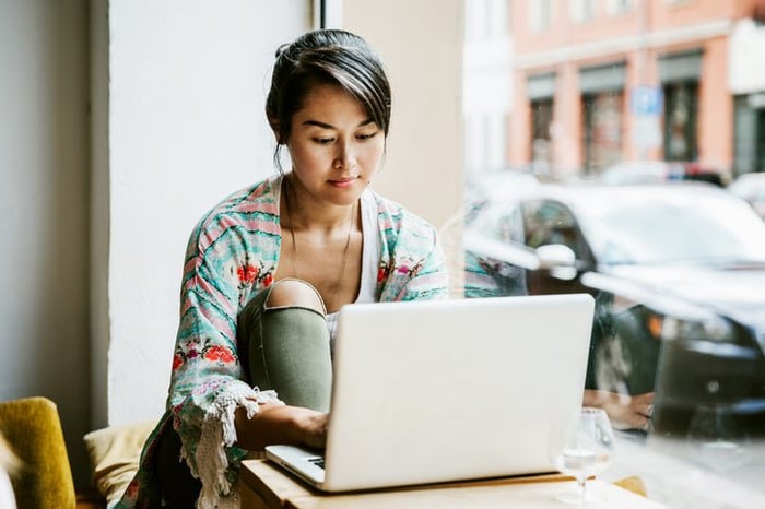 A woman sitting at a table in a cafe window and typing on a laptop.