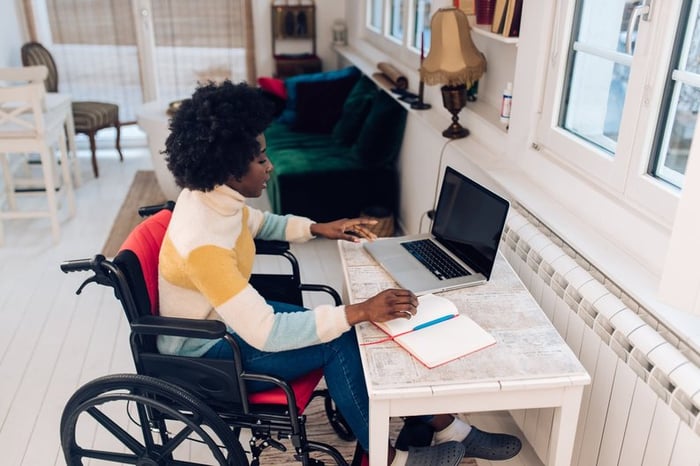 A person using a wheelchair types on a laptop at a desk at home.