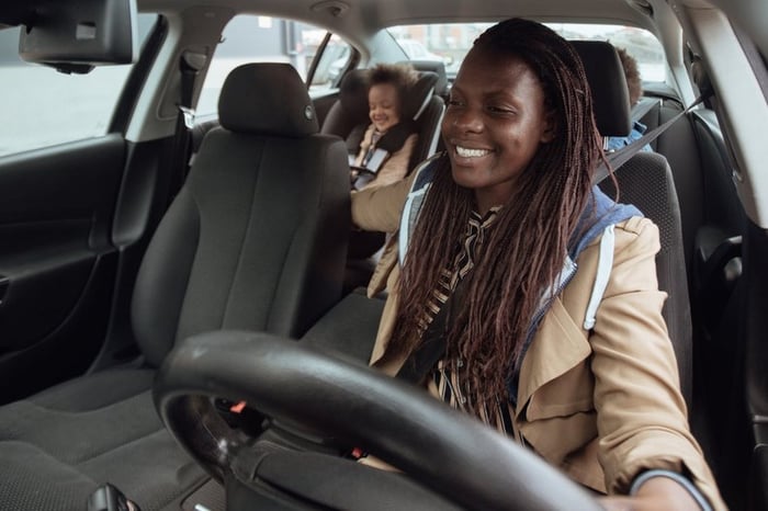 A smiling parent driving a car with their two kids in the backseat.