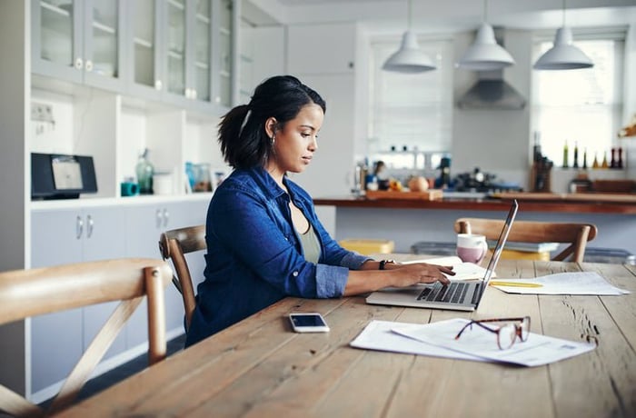 A woman sitting at her kitchen table and typing on a laptop next to paperwork.