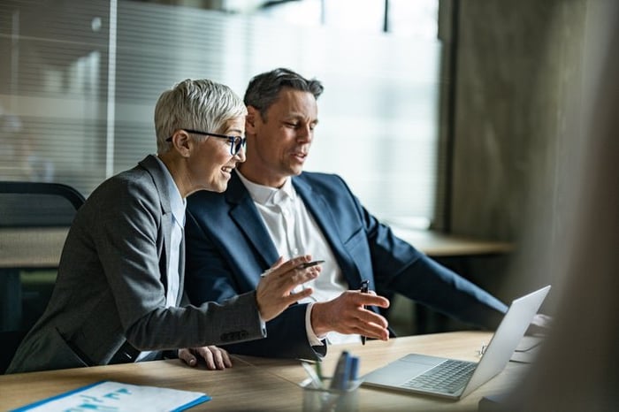 Two mature colleagues discuss while looking at a laptop in an office.