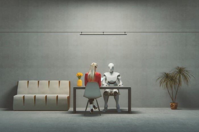 A person sitting at a desk across from a robot in a plain, gray room.