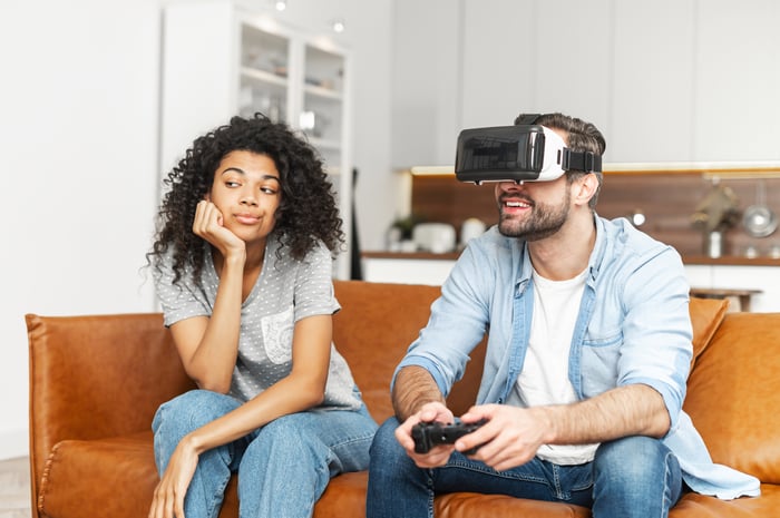 A couple sitting on a couch while one person wears a virtual realty headset and holds a game controller.