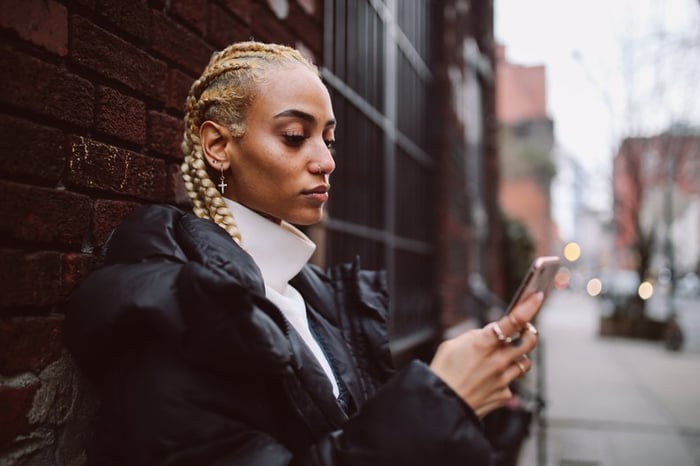 A young adult uses their phone while leaning against a brick wall in a city.