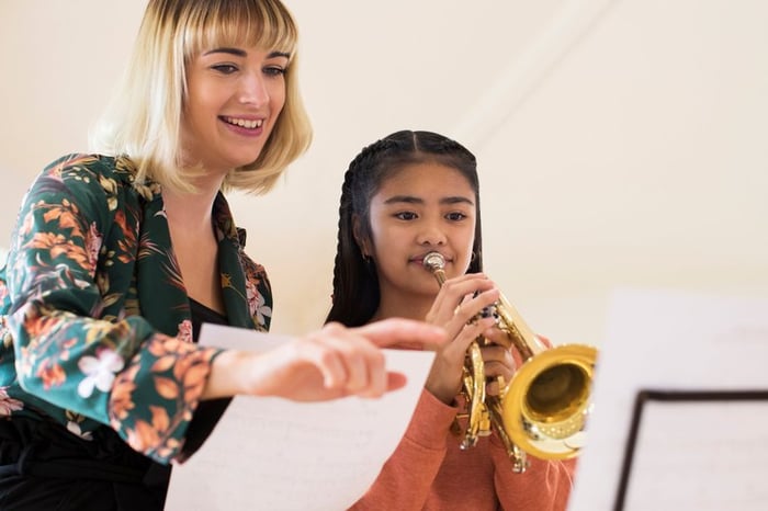 A smiling female teacher instructing a young girl playing a trumpet.