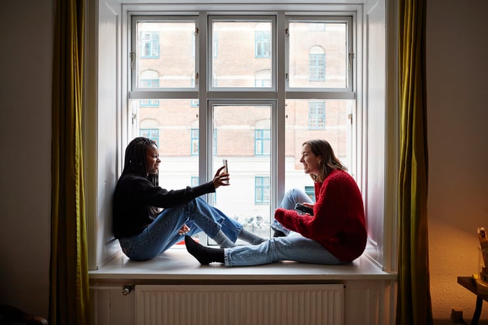 Two people sitting in a window alcove while one shows their phone screen to the other.