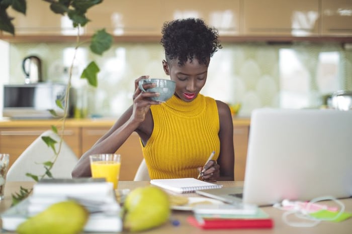 A woman drinking coffee at her kitchen table while writing in a notebook with her laptop open in front of her.