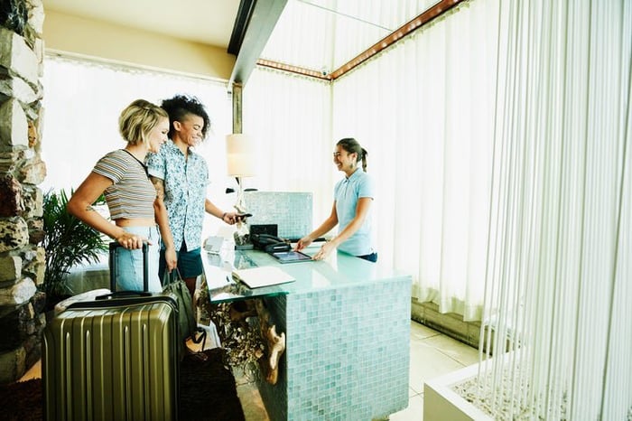 Two people with suitcases checking into a hotel by swiping their phone on the payment reader with an employee behind the lobby desk.