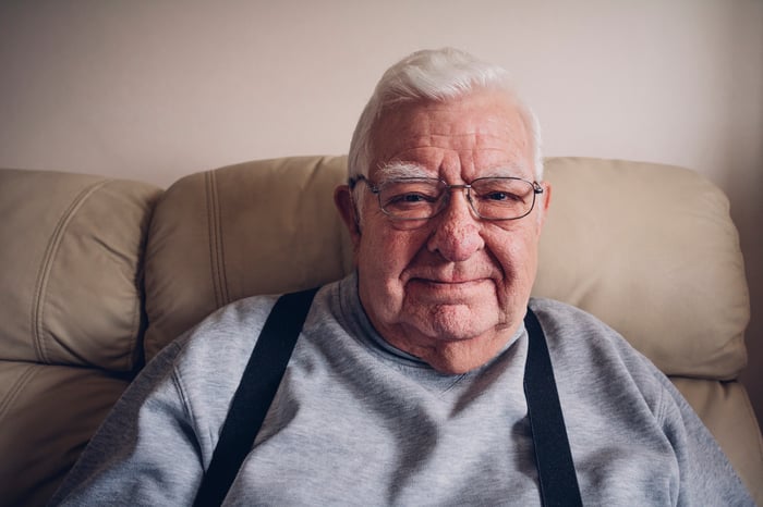 A senior citizen sitting on a couch, wearing suspenders, is smiling.