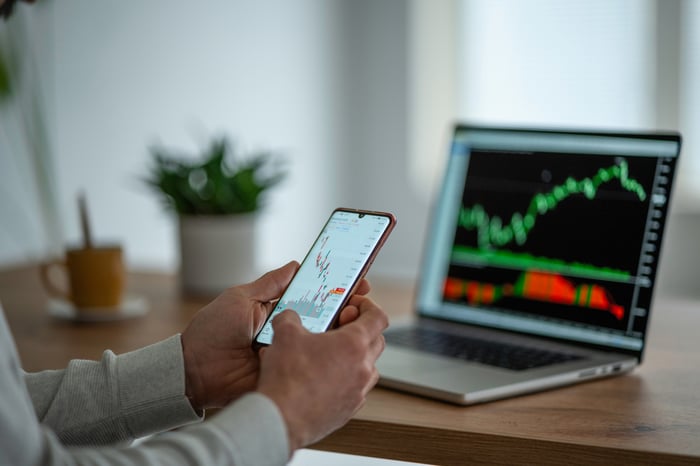 A pair of hands holds a smartphone displaying a graph in front of a laptop displaying another graph.