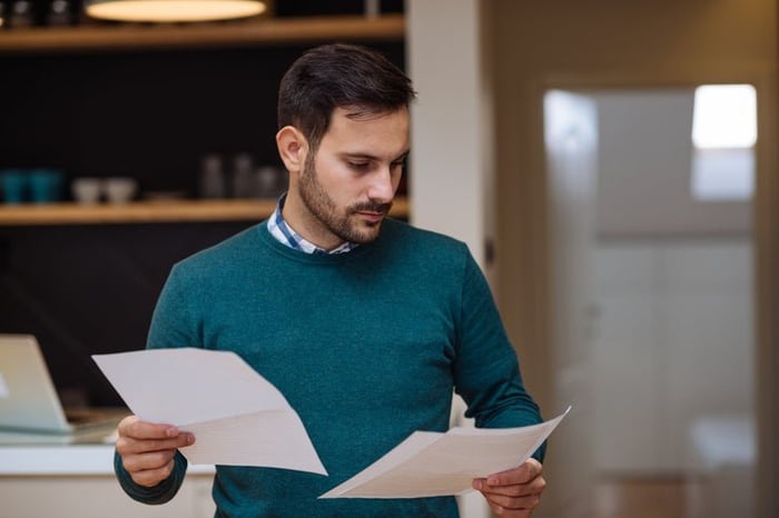 A man standing in his living room and looking between two pieces of paper held in each hand.