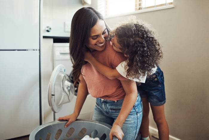 A kid hanging on to an adult carrying a laundry basket.