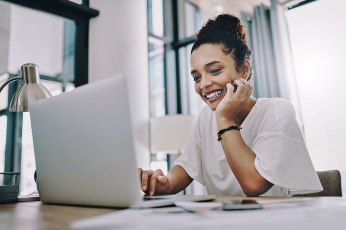 An investor in an office smiles while looking at something on a laptop.