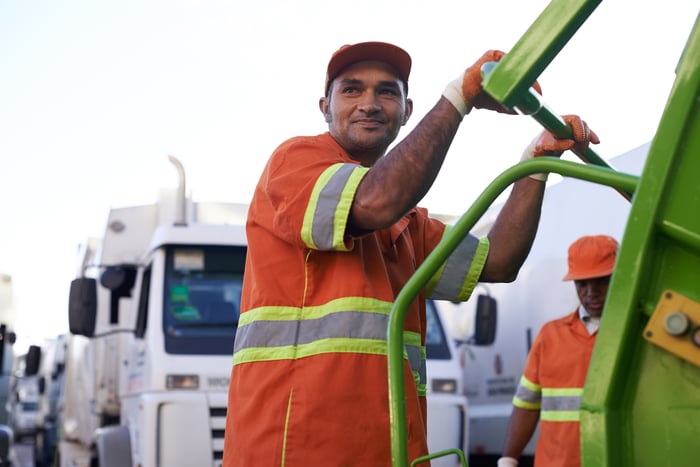 A person smiles while riding on a garbage truck.