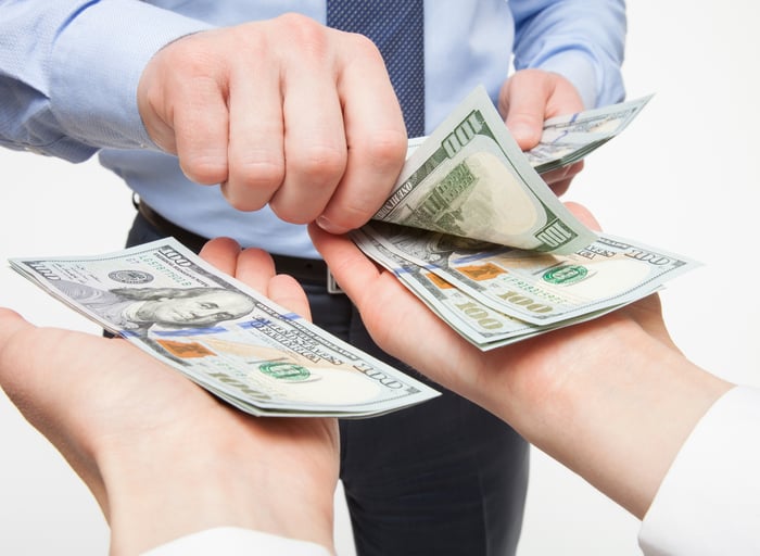 A businessperson placing crisp one-hundred-dollar bills into two outstretched hands. 