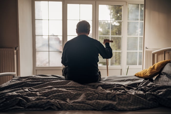 Person holding a cane sitting on the edge of a bed.