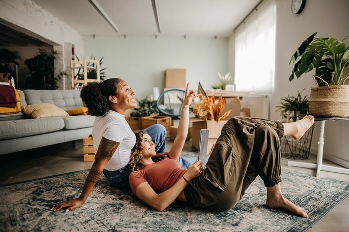 A couple smiles looking at a tablet while sitting on the floor of their apartment.