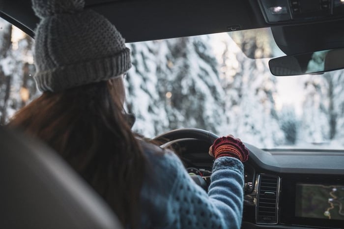 Woman in cold weather accesories driving a vehicle through snowy landscape.